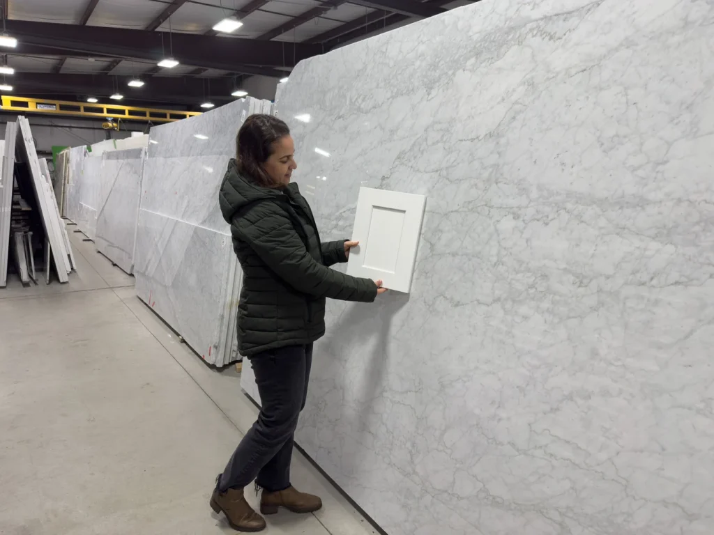 A young woman holds a white shaker cabinet panel up to a white Quartzite slab to see if they look good together